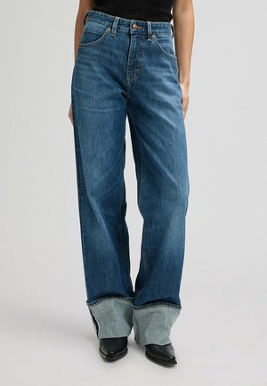 Kobieta w luźnych niebieskich dżinsach z dużymi podwiniętymi mankietami, nosząca czarne, spiczaste buty na tle jednolitego tła.