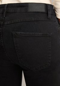 Czarne dżinsy o gładkiej fakturze, z tylną kieszenią i prostokątną skórzaną metką z wytłoczonym logo marki na pasku.