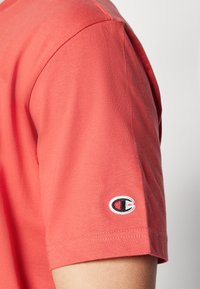 T-shirt en coton corail avec un design à manches courtes, orné d'un logo brodé blanc et noir sur la manche gauche. Texture lisse, coupe classique.