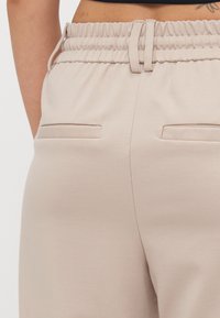 Jogging en tissu beige avec une taille élastique, comportant deux poches arrière avec des bords cousus et une texture lisse et douce.