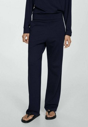 Persoon draagt losse donkerblauwe broek, bijpassende top met lange mouwen en zwarte teenslippers, staande tegen een eenvoudige lichte achtergrond.