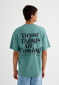 Camiseta de algodón color añil con gran texto en negro en la parte posterior: "¡Grandes cosas están por venir!" con texto adicional debajo. Mangas cortas, corte relajado.