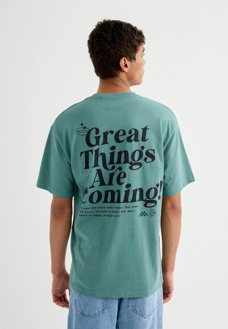 Camiseta de algodón color añil con gran texto en negro en la parte posterior: "¡Grandes cosas están por venir!" con texto adicional debajo. Mangas cortas, corte relajado.