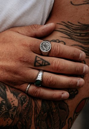 Three rings on a hand: a silver signet with a wave pattern, a textured silver band with a green stone, and a thin silver ring.