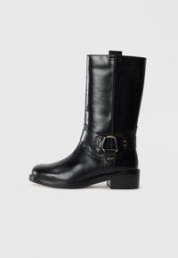 Botte en cuir noir, montant jusqu'au genou, avec un bout arrondi, une fermeture éclair sur le côté et une sangle décorative avec des accessoires en laiton et des clous. Talon plat.