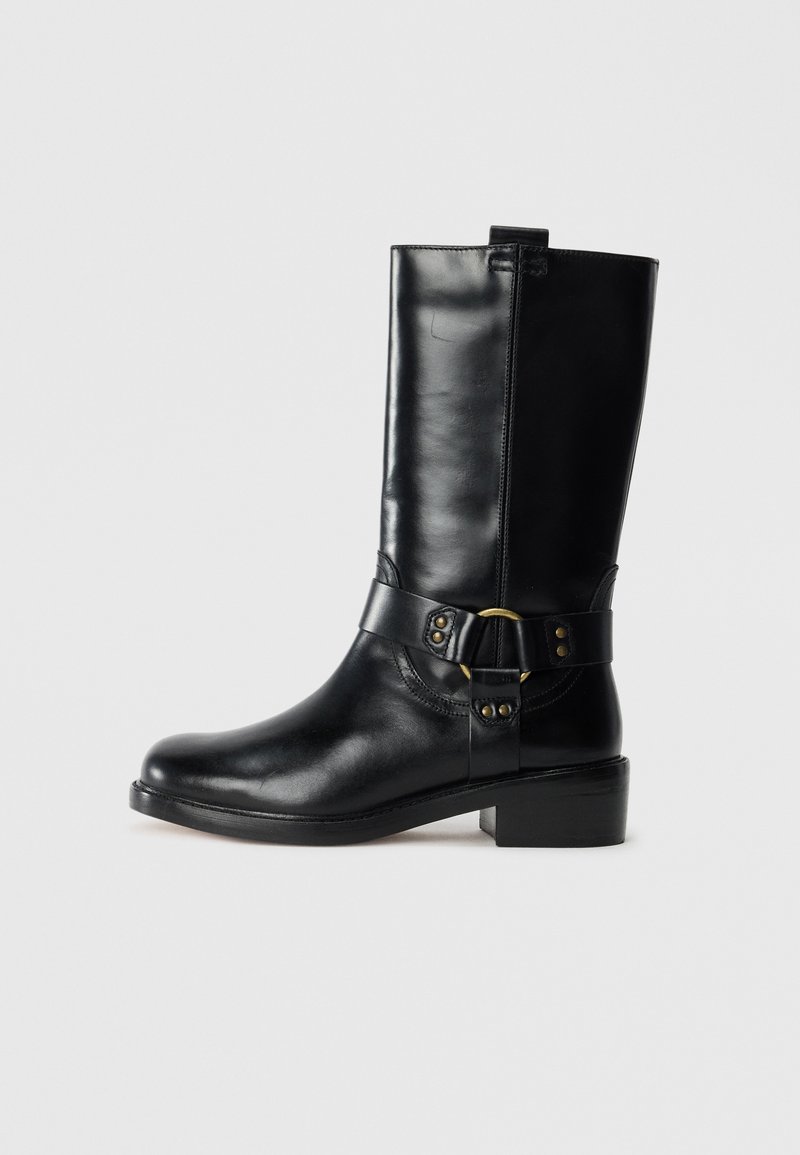 Botte en cuir noir, montant jusqu'au genou, avec un bout arrondi, une fermeture éclair sur le côté et une sangle décorative avec des accessoires en laiton et des clous. Talon plat.