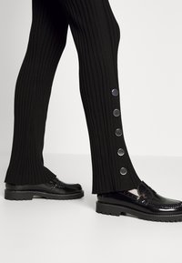 Svarta ribbade byxor med trumpetdesign, prydda med silverfärgade tryckknappar längs sidan. Matchat med blanka svarta loafers.