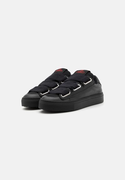 Sorte sneakers med et struktureret stofoverdel, brede sorte remme, sølv hardware-løkker og en flad gummisål. Rød logoakcent på tungen.