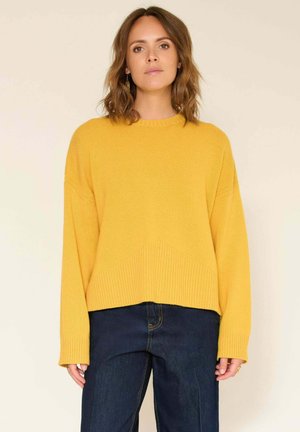 Pull en tricot jaune avec un col rond, des poignets côtelés et une coupe courte. Associé à un jean en denim bleu foncé. Texture lisse.