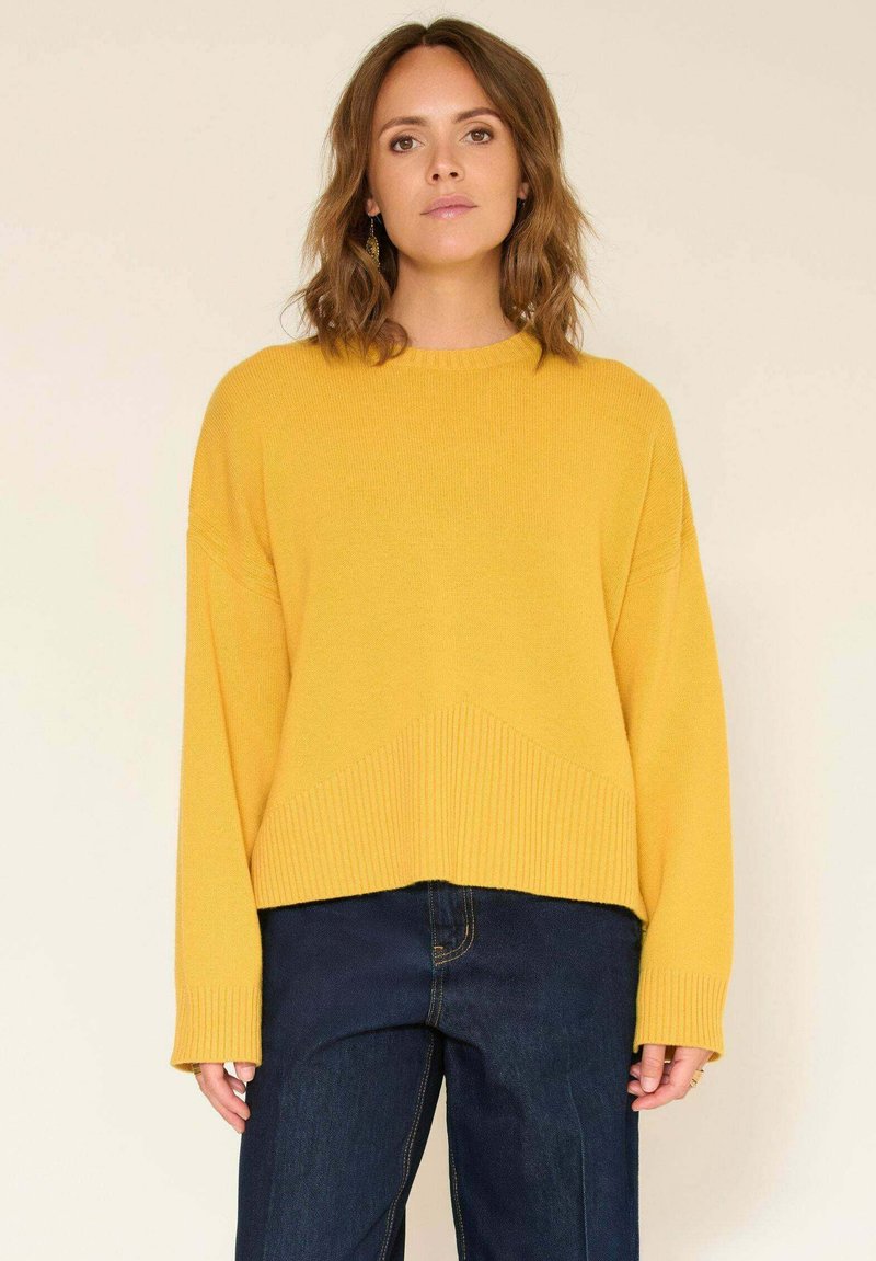 Pull en tricot jaune avec un col rond, des poignets côtelés et une coupe courte. Associé à un jean en denim bleu foncé. Texture lisse.