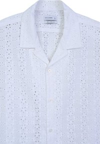 Camicia bianca a maniche corte con vestibilità rilassata, dotata di ricami ad occhio, bottone frontale e colletto classico.