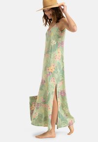 Lungo vestito floreale verde con fiori rosa e gialli, spacchi laterali e spalline regolabili; indossato con un cappello a tesa larga color beige.