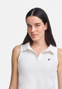 Polo shirt bianco senza maniche con una texture a coste, dotato di colletto e un piccolo logo nero sul lato sinistro.