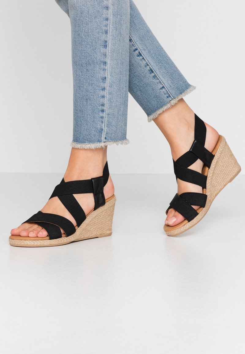 Pieds chaussés de jeans bleu clair à ourlet effilé et de sandales compensées noires à brides avec talons tissés beige sur un sol blanc.