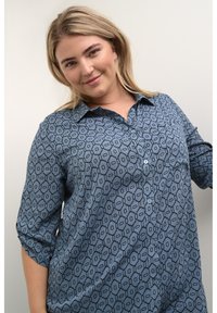 Niebieska koszula w wzory z guzikami z przodu, z możliwością podwinięcia rękawów oraz geometrycznym wzorem w ciemniejszym odcieniu niebieskiego. Miękka tkanina o luźnym kroju.