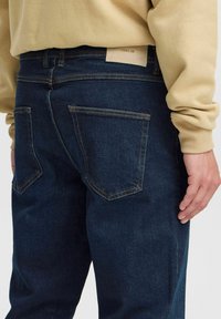 Jeans de mezclilla oscura con un corte clásico, con costuras en tono marrón y dos bolsillos traseros. Una etiqueta beige es visible en la cinturilla.