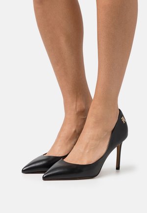 Ben klædt i sorte spidse læder højhælede sko med stiletto hæle, stående på hvid baggrund.