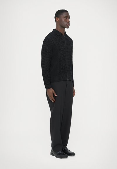 Pull noir en tricot avec un motif torsadé et un col, associé à un pantalon sombre et des chaussures noires. Le mannequin est de profil sur un fond blanc.