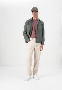 Cămașă verde cu nasturi deasupra unei tricouri vișinii, pantaloni crem și adidași albi. Pălăria are un model în carouri cu o margine neagră.