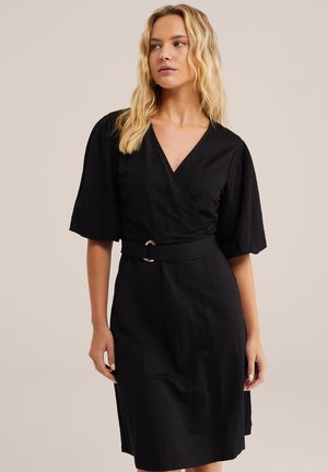 Lahtiste laineliste blondide juustega naine, kes kannab musta ümbermähitavat kleiti laiate varrukatega ja vööga, vaadates küljele tavalise tausta ees.