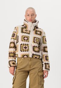 Fleece zip-up-jacka med diamantmönster i beige, rosa, grön och brun. Har en hög krage och texturerad tyg. Matchas med beiga cargobyxor.