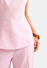 Γυναίκα που φοράει αμάνικη μπλούζα ανοιχτού ροζ και παντελόνι στο ίδιο χρώμα, με βραχιόλι κεχριμπαρένιου χρώματος στον δεξί καρπό, σε λευκό φόντο.