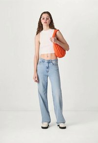 Kvinna i vit ärmlös crop top, ljusblå vida jeans, vita skor, vita solglasögon, som håller en orange axelväska framför en enfärgad bakgrund.