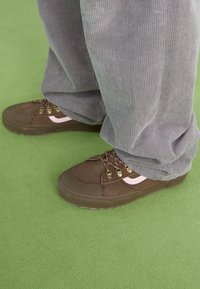 Zapatillas marrones de suède y goma, con acentos beige, cordones en contraste y puntera redondeada, llevadas con pantalones de pana grises.