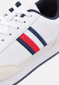 Sneaker bianca con tomaia in pelle liscia, strisce blu e rosse, dettagli in suede, lacci piatti bianchi e suola in gomma.