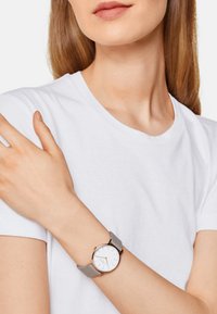 Runde Uhr mit einem weißen Ziffernblatt, rose-goldenen Details und einem hellgrauen Armband. Die Uhr wird an einem hellhäutigen Arm getragen. Ein weißes T-Shirt ist sichtbar.