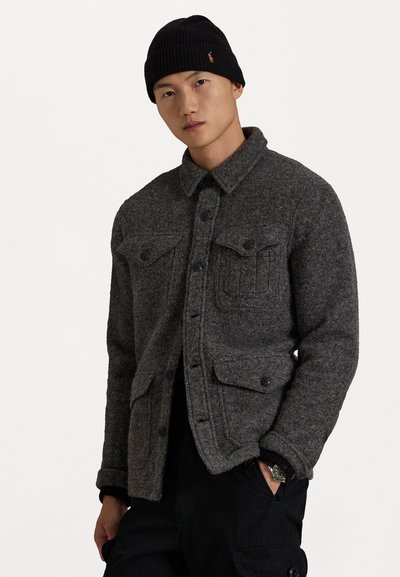 Grå uldsjakke skjorte med brystlommer, knaplukning og sort ribbet hue. Slim fit og struktureret stof. Sorte bukser delvist synlige.