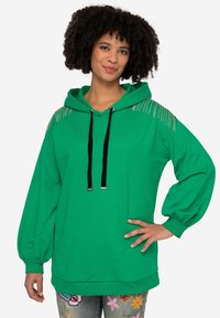 Felpa verde realizzata in morbido tessuto, con cappuccio, lacci neri e dettagli metallici sulle spalle. Maniche ampie e vestibilità rilassata.