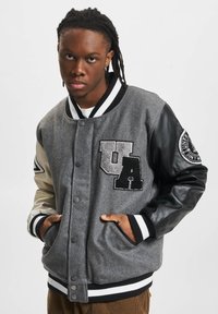 Grå ullblandings varsity-jakke med svarte skinnerm, U.A.-lapp på brystet, stripete mansjetter og skjulte sidelommer.