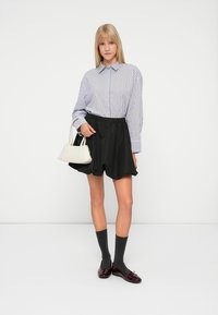 Wit en blauwe gestreepte langemouwen shirt, zwarte geplooide shorts, grijze sokken, bordeauxrode schoenen en een kleine witte handtas. Effen achtergrond.