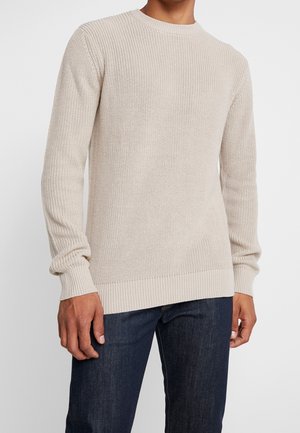 Béžový žebrovaný svetr vyrobený z texturované pleteniny, s kulatým výstřihem a dlouhými rukávy, v kombinaci s tmavě modrými džíny.