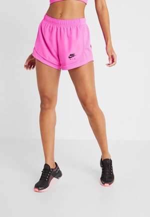 Vrouw die felroze Nike Air sportshorts en zwarte hardloopschoenen draagt, staand tegen een effen lichte achtergrond.