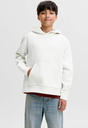 Garçon portant un sweat à capuche blanc, une main dans la poche avant et un jean bleu clair, debout devant un fond gris uni.