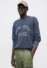 Granatowy sweatshirt z okrągłym dekoltem z napisem "NOWY JORK" w kolorze białym, w zestawieniu z oliwkowymi spodniami cargo. Gładki bawełniany materiał, luźny krój.