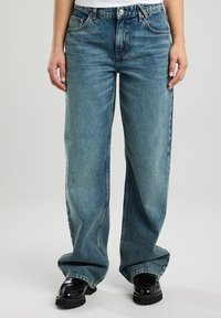Verbleekte blauwe denim wijde broek met hoge taille, voorzien van voorzakken en een knoopsluiting. Zwarte chunky schoenen maken de outfit compleet.