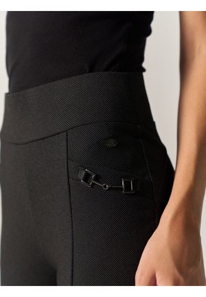 Plano detalle de una persona que lleva pantalones negros de cintura alta con textura y un pequeño detalle de hebilla plateada cerca de la cadera y un top negro.