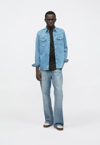 Ανοιχτό μπλε τζιν πουκάμισο με κουμπωτή μπροστινή πλευρά και δύο τσέπες στο στήθος, φοριέται πάνω από μαύρο πουκάμισο. Συνδυάζεται με ανοιχτό μπλε φαρδύ τζιν και αθλητικά παπούτσια.