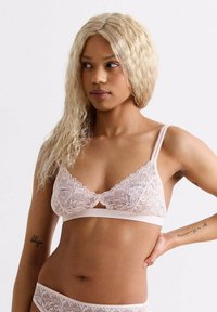 Lace bralette in lichtroze met verstelbare bandjes, bloemenpatroon en een sleutelgatdetail in het midden. Zacht materiaal, minimale hardware.