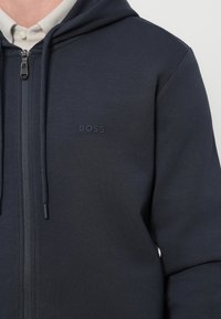 Námornícka mikina na zips vyrobená z mäkkej tkaniny, s kapucňou na sťahovanie, rebrovanými manžetami a malým, vyrazeným logom "BOSS" na hrudi.