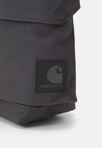 Sort stofpose med klaplukning, udstyret med en rund trykknap og en firkantet lap med et logo. Holdbar tekstur med stikninger.