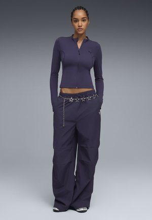 Vrouw draagt een donkerpaarse lange mouwen top met ritssluiting en wijd uitlopende broek met een zilveren sterrenkettingriem, staand met handen in de zakken.