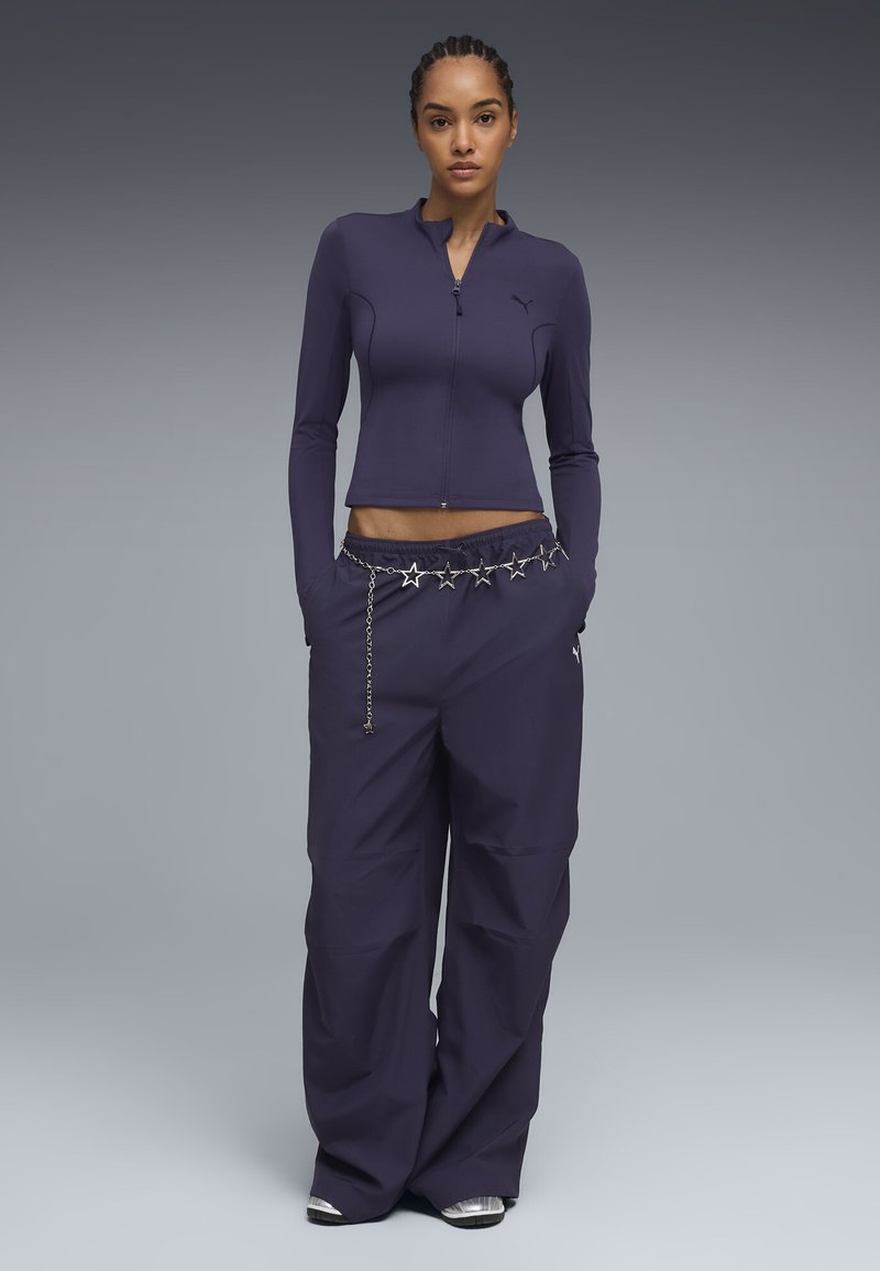 Femme portant un haut à manches longues zippé violet foncé et un pantalon large avec une ceinture chaîne étoile argentée, debout les mains dans les poches.
