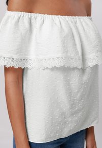 Weißes Off-Shoulder-Oberteil aus texturiertem Stoff mit besticktem Spitzensaum am Ausschnitt, das ein gepunktetes Muster aufweist.