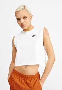 Camiseta sin mangas blanca de corte crop en algodón con cuello redondo y logotipo negro de Nike en el pecho izquierdo. Combinada con pantalones naranjas.