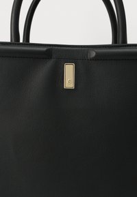 Μαύρη δερμάτινη τσάντα χειρός με δύο λαβές και μικρό ορθογώνιο χρυσό έμβλημα στο κέντρο κοντά στην επάνω άκρη.