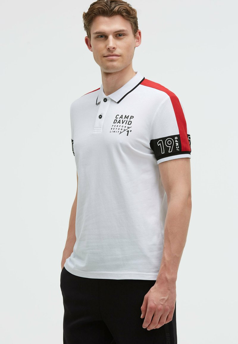 Polo bianco con accenti rossi e neri sulle spalle, caratterizzato da una banda nera con "19 CMPD." Include un colletto con bottoni e testo stampato.